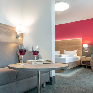 Im Doppelzimmer Komfort Plus stehen zwei Weingl&auml;ser auf einem kleinen Tisch und im Hintergrund ist der Schlafraum zu erkennen