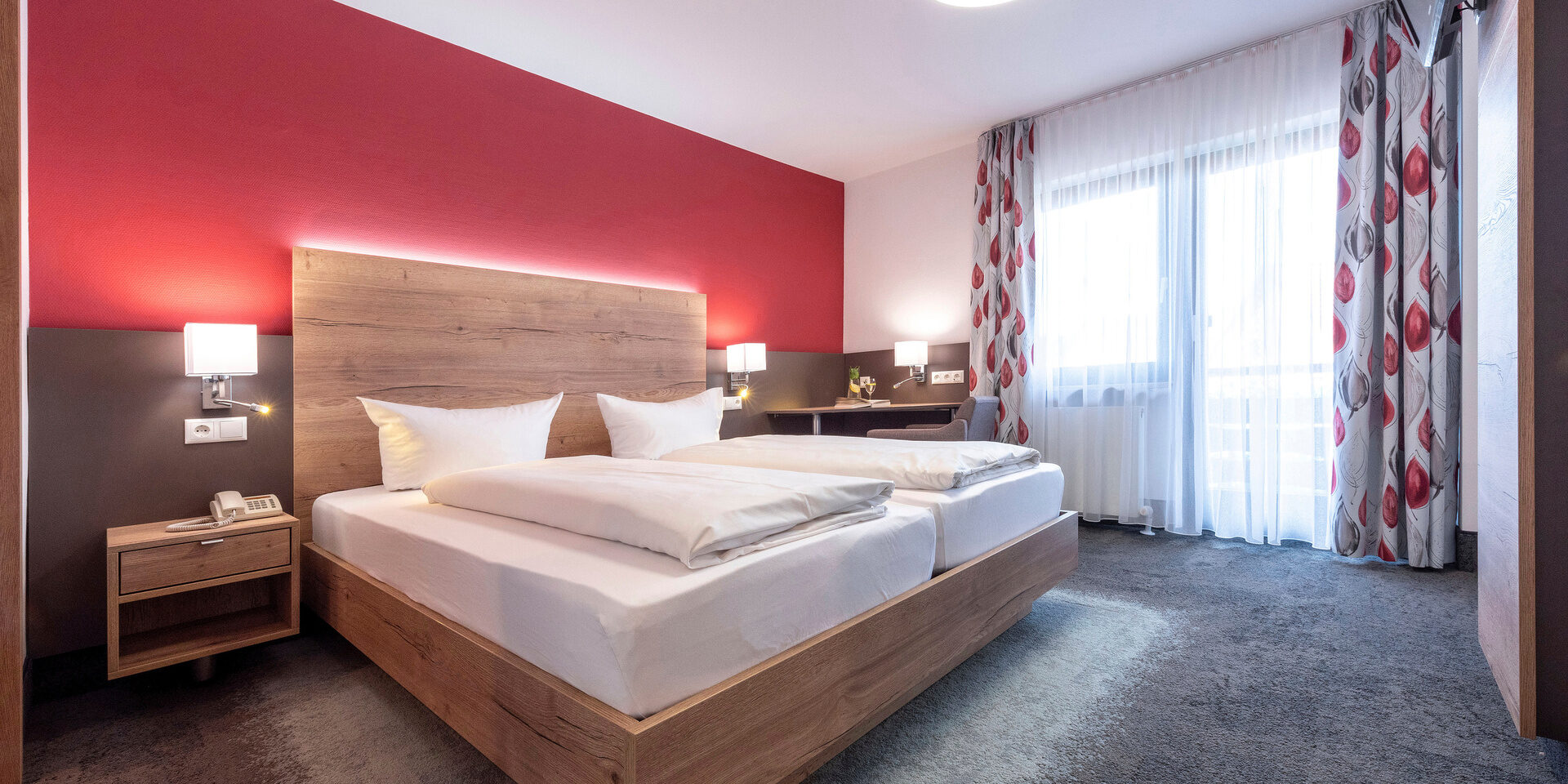 An Kopfende des Betts im Doppelzimmer Komfort ist eine LED-Leiste Verbaut und die rote Wand und das Holz des Bettes hervorzuheben.