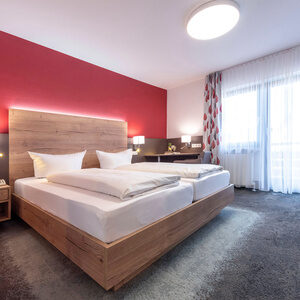 An Kopfende des Betts im Doppelzimmer Komfort ist eine LED-Leiste Verbaut und die rote Wand und das Holz des Bettes hervorzuheben.